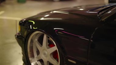 Parlak, siyah, klasik bir arabanın gece otoparkta yavaşça sürüşünün yakın çekimi. Manzara gümüş alaşımlı tekerlek, kırmızı fren kalibreleri ve koyu renk boyaya odaklanıyor.