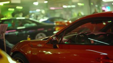 Bir motor sergisinde canlı yeşil ve mor neon ışıkların altında parlayan modern kırmızı spor araba. Diğer özelleştirilmiş araç ve izleyiciler arka planda bulanıklaşarak heyecan verici bir atmosfer yaratıyor.