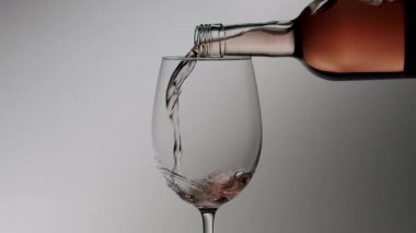 Temiz cam şişe zarif bir kristal bardağa pembe gül şarabı döküyor, damlalar ve kabarcıklar yavaş çekimde, temiz, sade ve nötr bir arka plana karşı.