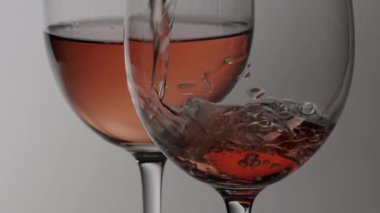 Temiz cam şişe zarif bir kristal bardağa pembe gül şarabı döküyor, damlalar ve kabarcıklar yavaş çekimde, temiz, sade ve nötr bir arka plana karşı.