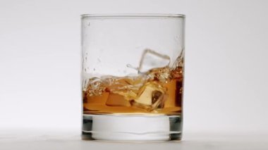 Kristal berrak buz küplerinin altın kahverengiyle dolu bir bardağa düşüşü çarpıcı bir sıçrama ve girdap yaratıyor, temiz beyaz bir arkaplanda izole edilmiş.