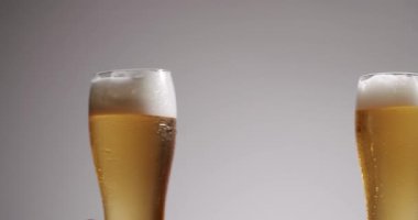 Gri bir arka planda, iki kişi bir bardak taze altın birayla kutlama ve kadeh kaldırıyor. Soğuk bira tokuşturarak yavaş çekimde bir sıçrama ve köpük oluşturur.