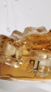 Kristal berrak buz küplerinin altın kahverengiyle dolu bir bardağa düşüşünü gösteren yavaş çekim videosu temiz beyaz bir arkaplanda dramatik bir sıçrama ve girdap yaratıyor. Dikey