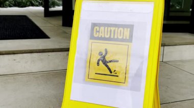 Bir binanın girişindeki fayanslı zeminde duran sarı uyarı levhası insanları kazaları ve yaralanmaları önlemek için ıslak ve kaygan bir yüzey tehlikesi konusunda uyarıyor.