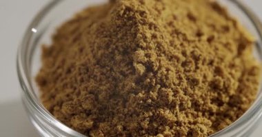 Şeffaf bir cam kasede şeffaf bir zencefil tozu, baharat dokusunu ve zengin kahverengi rengini, ayrıntılı bir makro çekimde temiz beyaz arka plana karşı yavaşça gösteriyor.