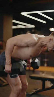 Modern bir spor salonunda kaslı bir adam sırayla yatıyor. Gömleksiz ve odaklanmış, halterleri kaldırırken sırt kaslarını gösteriyor. Bu video formda güç, güç ve kararlılığı yakalar.