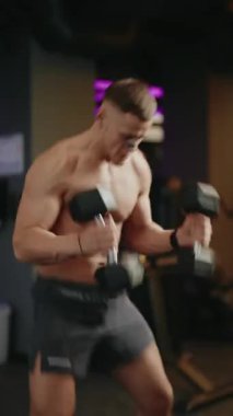 Kas yığını bir adam spor salonunda dambıl itici egzersizi yapıyor. Dambılları yerden havaya kaldırıyor, dayanıklılık ve zindelik gösteriyor. Spor salonu ayarları onun eğitime olan bağlılığını vurguluyor.