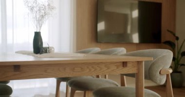 Güneşli modern yemek odasında, içinde kurumuş çiçeklerden oluşan bir vazo, zarif sandalyeler, eğimli bir duvarda televizyon ve katıksız perdeleri olan büyük bir pencere olan ahşap bir masa, rahat bir atmosfer yaratıyor.