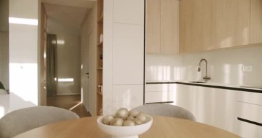 Güneşli sabah ışığı modern ve minimalist bir apartman dairesinde gölgeler oluşturuyor. Ahşap bir yemek masası, çağdaş mutfak dolapları ve parlak bir koridor manzarası var.