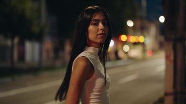 Çekici esmer bir kadının yavaş çekimde portresi gecenin bir yarısı boş bir şehir sokağında kameraya doğru dönüyor, bulanık araba ışıkları arka planda güzel bir bokeh etkisi yaratıyor.