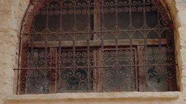Eski, yıpranmış, karmaşık işlenmiş demir ızgaralı pencerenin yakın görüntüsü. Pencere, geleneksel Fas mimarisinin tipik bir örneği olan pürüzlü taş ya da sıva duvarına sabitlenmiş..