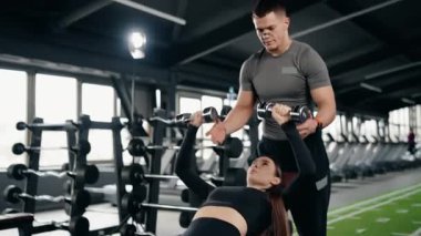 Genç bir bayana modern bir spor salonundaki eğimli bir bankta halterli göğüs egzersizi yapması için yardımcı olan beden eğitimi hocası, eğitim sırasında düzgün forma ve kas gelişimine odaklanıyor.
