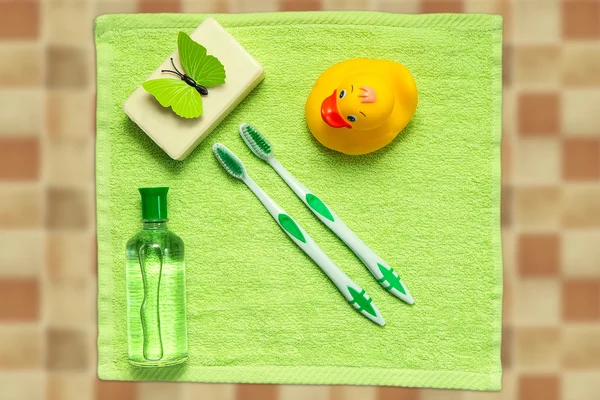 Accessoires de salle de bain sur une serviette verte. Canard en caoutchouc, brosses à dents, savon et lotion. Accessoires de soins pour bébé pour bain. Jouet de canard jaune pour enfants . — Photo