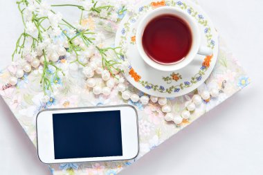 Bir fincan çay ve bir beyaz saten arka plan, bir kitap, çay bardağı, telefon ve dekorasyon çiçekli inci kolye ile telefon