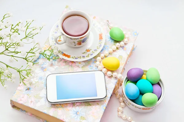 Phone with a cup of tea and pearl necklace with flowers on a white satin background. A book, tea cup, telephone and decoration. Tea time and easter painted eggs — Stock Photo, Image