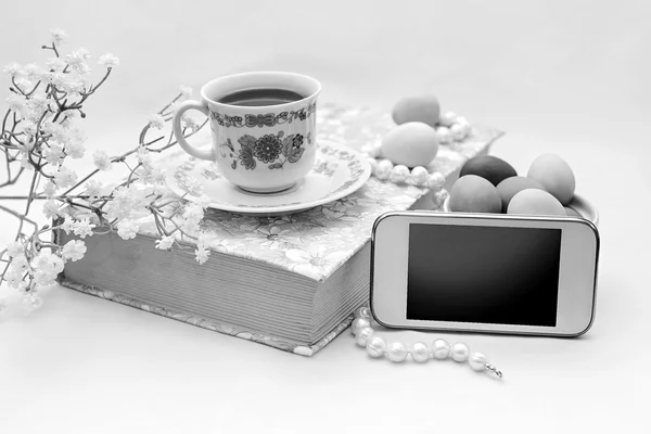 Phone with a cup of tea and pearl necklace with flowers on a white satin background. A book, tea cup, telephone and decoration.