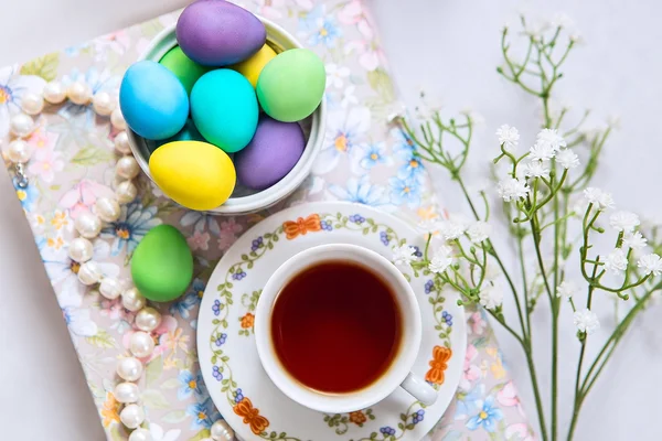 A cup of tea with easter painted eggs and some decorations - flowers, pearl necklace and a book. — Stock Photo, Image