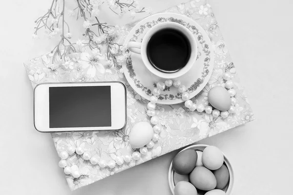 Phone with a cup of tea and pearl necklace with flowers on a white satin background. A book, tea cup, telephone and decoration. Tea time and easter painted eggs