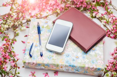 Ofis aksesuarları bir güzel çiçek dallarda beyaz bir arka plan ile. Telefon, defter ve bir kalem bir kitapla.