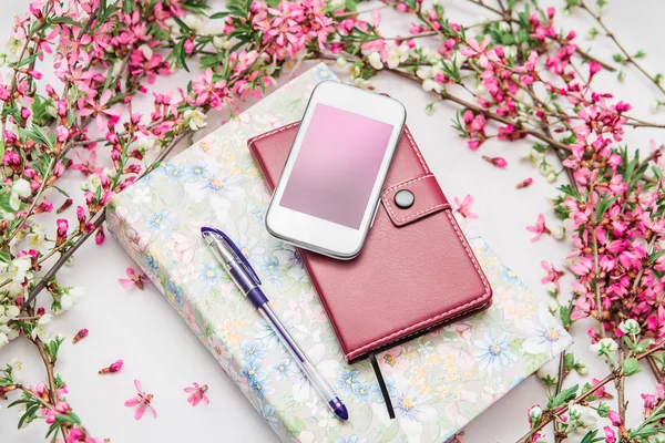 Office accessories with a beautiful blossom flower branches on a white background. Phone, notebook and a pen with a book.