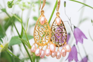 Pembe doğal inci ve altın işlemeli sarkık küpeler. Arka planda çiçek desenli zarif kadın aksesuarları.