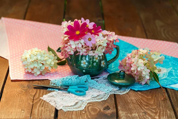 A bouquet of flowers in a teapot on a wooden table. Kitchen utensils, still life on a garden background. Plants, lace, polka dot fabric