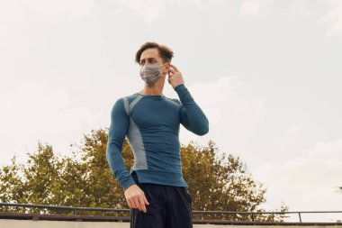 Spor kıyafetli, tıbbi maske takan genç bir adamın portresi. Covid-19 ölçümlerinden dolayı sarı tıbbi maske takan çekici bir erkek model. Beyaz adam kendini korumak için maske takıyor.