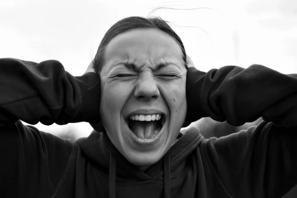 A young woman of 18-25 years old screams with an open mouth, covers her ears with her hands, refuses to listen, closes her eyes, frowns, showing her pain, express emotions, copy space.