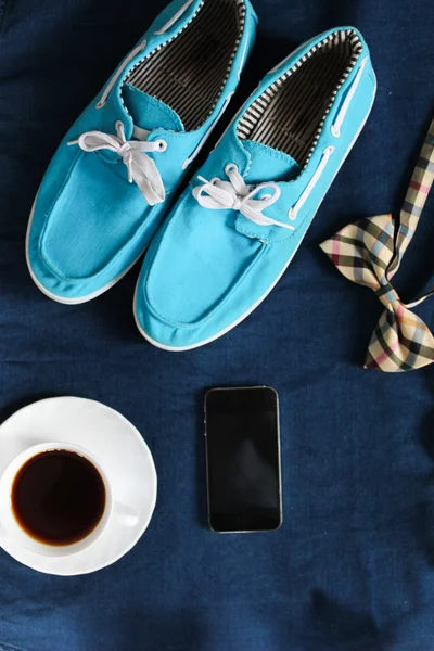 Top view of men`s casual shoes, mobile phone, cup of hot tea and bowtie on jeans background.