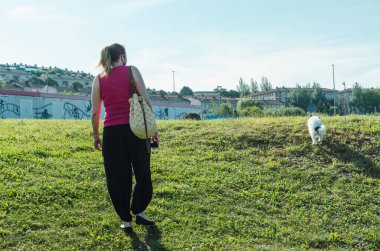 Parkta küçük köpekli genç bir kadın yürüyor.