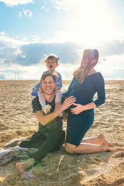 Üç kişilik aile kumda oturuyor ve gülüyor. Babası kızı omuzlarında ve annesi hamile. Mutlu hamilelikler ve ebeveynlik kavramı.