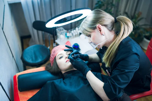 Permanent make up artist in black uniform and gloves holds tattoo machine upon her client lips with blurres lamp, a chair and equipment in the background. Skin care, permanent make up concept.