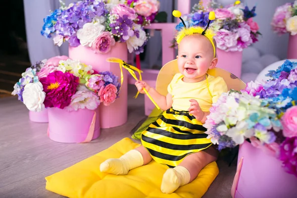 A smiling babygirl in a costume of a bee is sitting on a yellow pillow among bright flowers. She holds a bee on a stick.