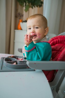 Kirli yüzlü bir oğlan çocuğu elleriyle yer. O, üzerinde tabağı olan beyaz masada yüksek bir sandalyede oturuyor. Sütten kesilme konsepti.