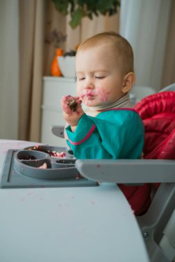 Kirli yüzlü bir bebek, elinde tuttuğu bir yiyecek parçasını inceliyor. O, üzerinde tabağı olan beyaz masada yüksek bir sandalyede oturuyor. Sütten kesilme konsepti.
