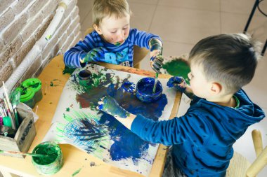 İki kardeş el ve fırçayla resim yapıyor, küçük çocuk odakta, diğeri ise bulanık. Ellerine boya bulaşmış ve resim çok dağınık..