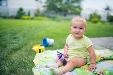 Bahçede yeşil bir battaniyenin üzerinde oturan sevimli bir erkek bebek. O, ciddi olarak yukarılara bakıyor. Arka planda büyük bir yeşil çimen alanı var..