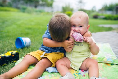 İki çocuk yeşil bahçede battaniyeye oturup birbirleriyle oynuyorlar. Büyük olan erkek kardeşine bakıyor. Arka planda yeşil çimenli bir alan var..