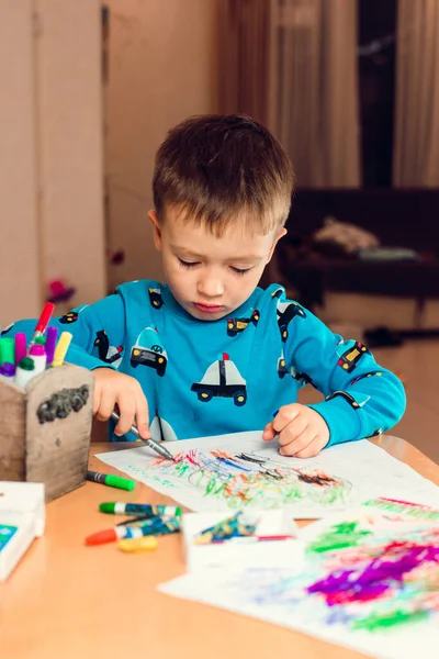 Tatlı bir çocuk beyaz kağıda suluboya kalemler ve pastel boyalarla parlak çizgiler çiziyor. Masada oturuyor ve pijama giyiyor..