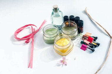 Kozmetik kavanozları, biberonlar, bitkisel kremler ve esans içeren şişelerle dolu bir hayat beyaz bir yüzeye serili pembe bir çiçek ve kağıt kurdelelerle. Doğal kozmetik konsepti.