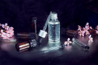 Gümüş kapaklı sade cam matara gümüş kapaklı cam damlatıcı ve siyah şapkalı, hepsi siyah arkaplana yerleştirilmiş su damlalarıyla kaplı. Etrafına kaos içinde bir sürü küçük düz matara ve pembe çiçekler yerleştirilmiş..