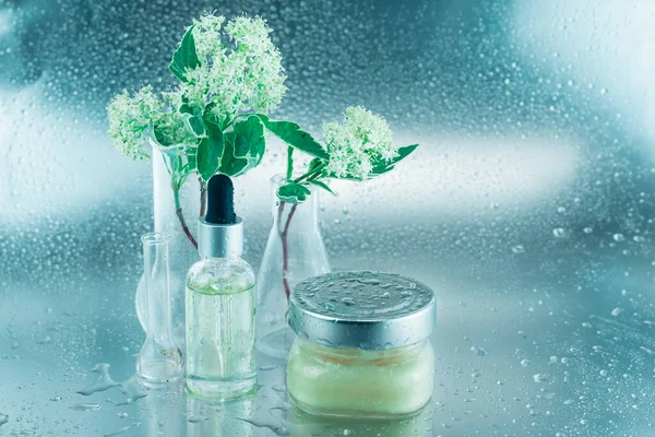 Glass flasks with flowers, a bottle of oil and big jar of cream on a silver background covered with drops. Natural cosmetical ingredients and essential oils concept.