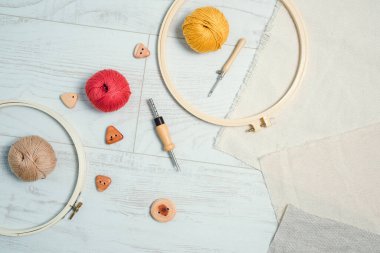 Embriyodery çemberlerin düzeni, yumruk iğneleri, iplik topları, keten kumaş ve tahta düğmeler beyaz ahşap zeminde. Yaratıcı işlemeler için araçlar ve ek malzemeler.