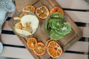 İki bardak kurutulmuş kivi ve yukarıdan çekilmiş elma. Ahşap tahta, arka planda kurutulmuş mandalinalar..