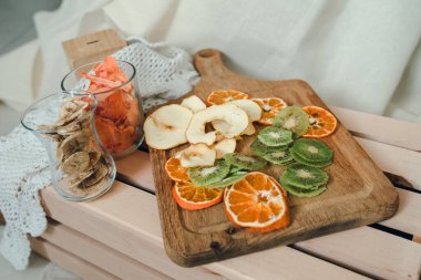 Kurumuş meyve düzeni - kivi, elma, mandalina, muz, balkabağı, bazıları ahşap tahtada, bazıları da buruşuk tuval arkaplamalı bardaklarda. Vegan sağlıklı tatlı konsepti.