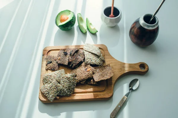 Ahşap tahtada servis edilen keten ekmek dizilimi. Arka planda taze avokado, sosis ve kabak var. Hepsi beyaz yüzeyde sert ışık huzmeleri var. Sağlıklı yemek..