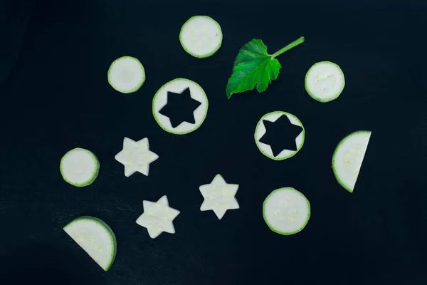 Slices of zucchini cutted in shapes of circles, stars and crescents and green zucchini leaf layed on dark black bachground. Healthy vegan food concept.