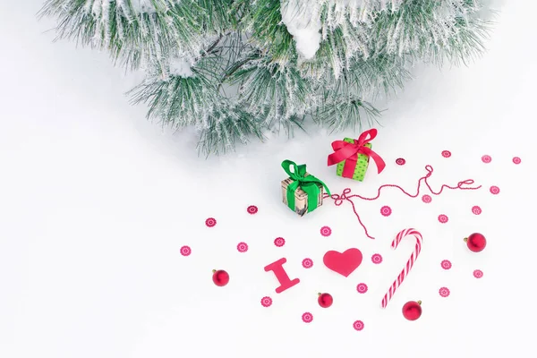 Kırmızı renkli Noel dekoratif elementlerinin üst görüntüsü - konfeti, hediye kutuları, boncuk şeridi, toplar, sert şeker, tabelayı seviyorum, hepsi köknar dallarının yanındaki beyaz yüzeyde, karla kaplı ve hoar.