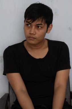 Bir adam kapalı bir yerde kamerayla doğrudan göz teması kurarken, siyah bir gömlek giyerken ve rahat bir şekilde otururken görülüyor. Tarafsız arka plan yüz ifadesini ve ruh halini vurguluyor. Uygun. 