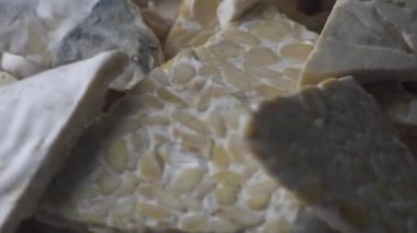 Son derece yakın plan çekimi, ham tempeh (mayalanmış soya fasulyesi) ve taze üçgen tofu dilimlerinin dokusunu gösterir. Bu dinamik görünüm bu anahtarın kalitesini ve bileşimini vurgular